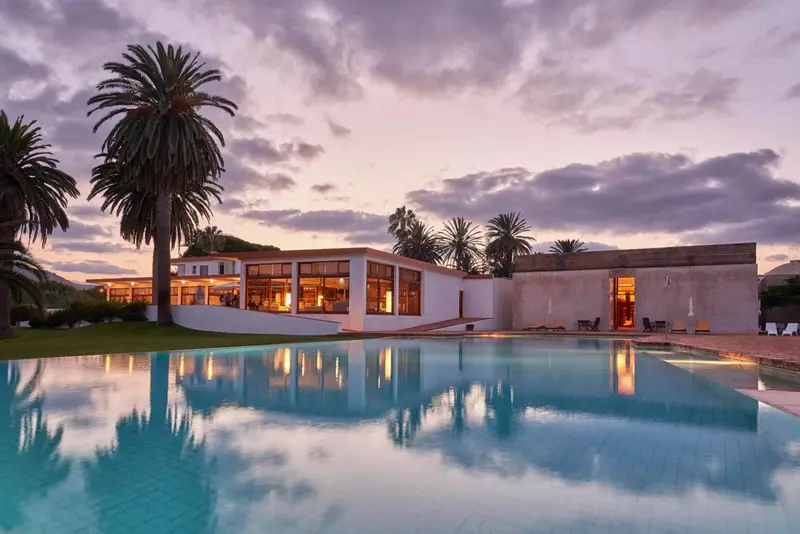 Bâtiment moderne avec piscine au premier plan et palmiers au coucher du soleil.