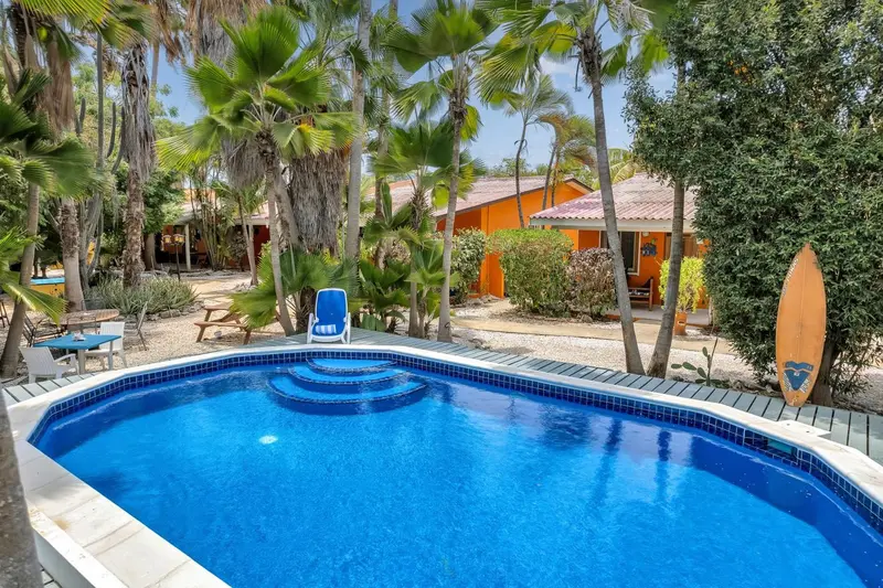 Piscine bleue entourée de végétation tropicale et de bâtiments colorés.