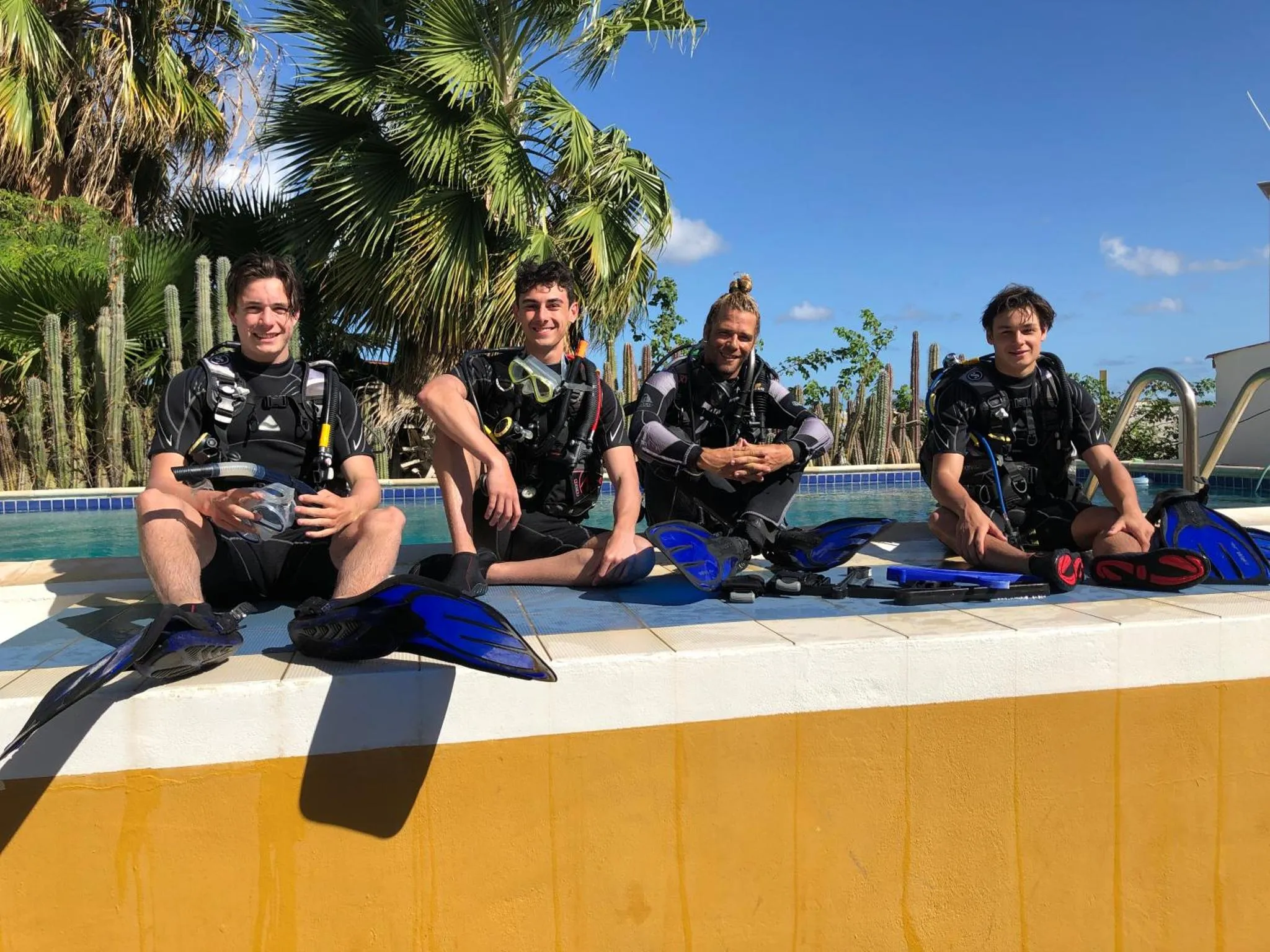 Quatre personnes assises au bord d'une piscine avec des palmes et des équipements de plongée.