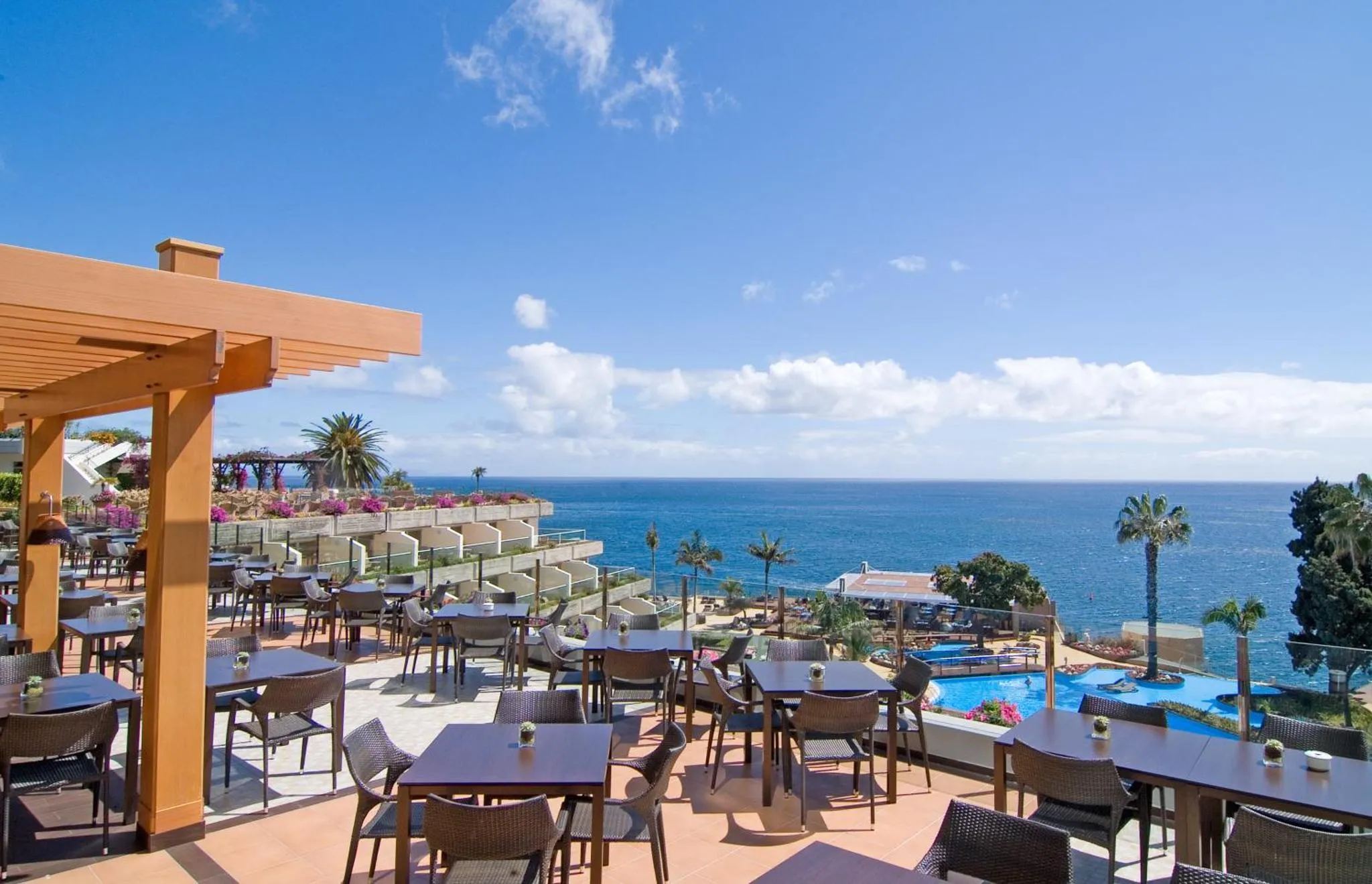 Terrasse ombragée avec tables et chaises donnant sur la mer et une piscine.