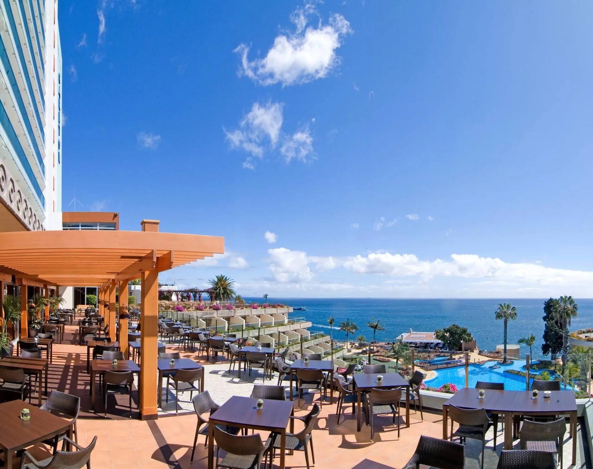 Terrasse ombragée avec tables et chaises donnant sur la mer et une piscine.
