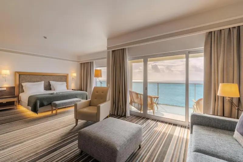 Chambre lumineuse avec un grand lit et un accès à un balcon donnant sur la mer.