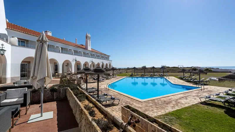 Une piscine rectangulaire bordée de transats et de parasols devant un bâtiment blanc à deux étages.