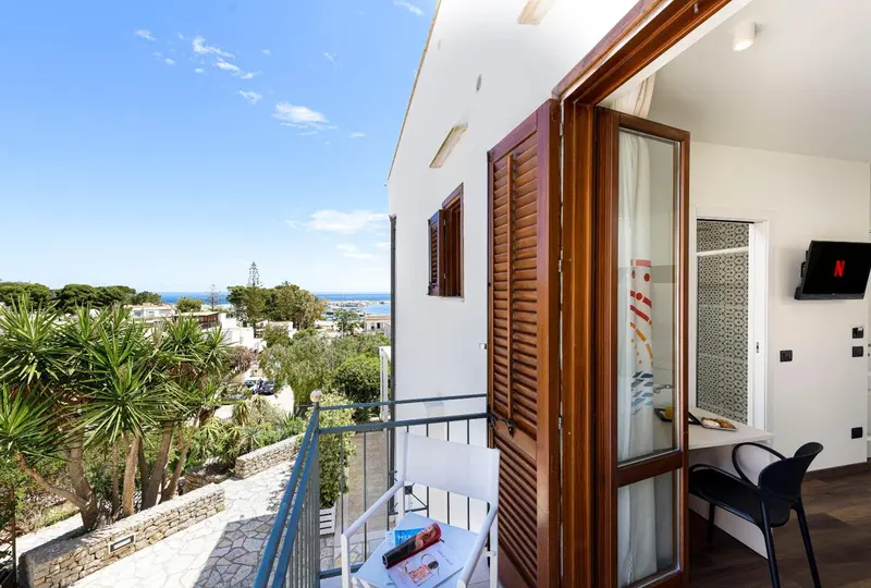 Une chambre lumineuse s'ouvre sur un balcon avec vue sur la végétation et la mer.