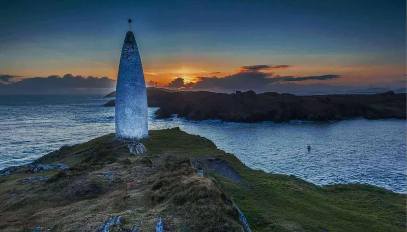 Un monument élancé se dresse sur une côte rocheuse au coucher du soleil.