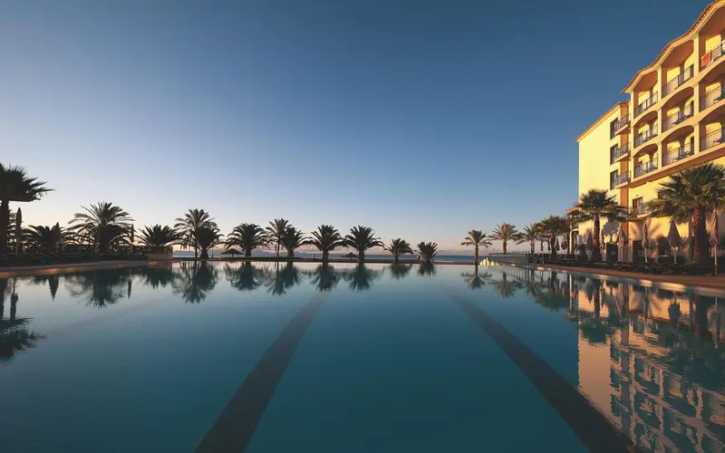 Une piscine rectangulaire reflète un bâtiment et des palmiers sous un ciel bleu.