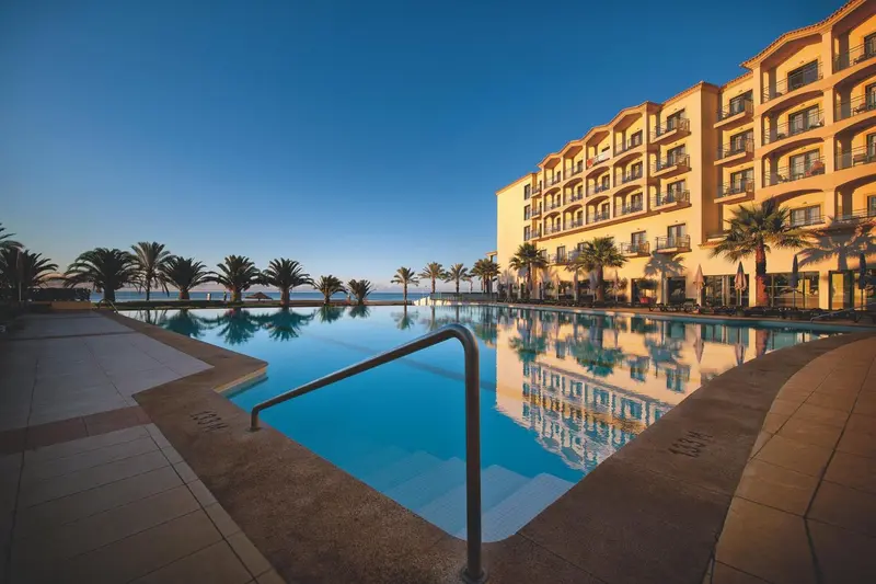 Une piscine rectangulaire reflète un bâtiment beige illuminé au crépuscule.