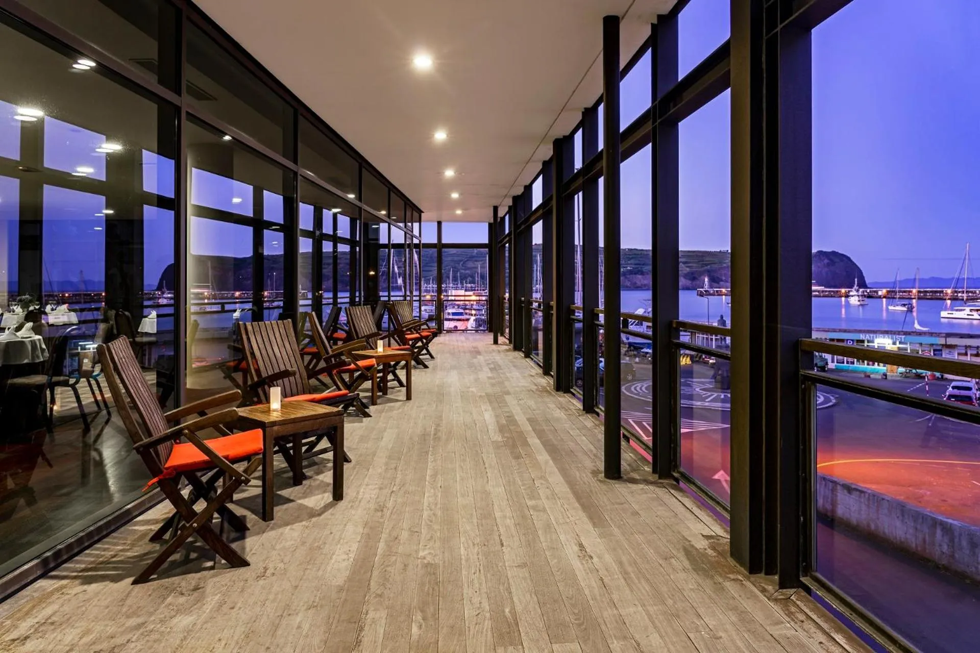 Terrasse avec mobilier en bois donnant sur un port illuminé au crépuscule.