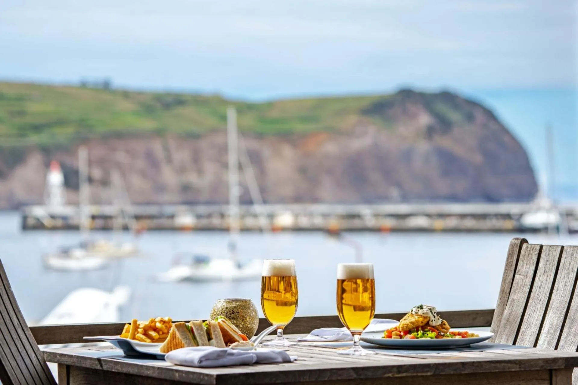 Une table est dressée avec des plats et des boissons en terrasse avec vue sur un port.