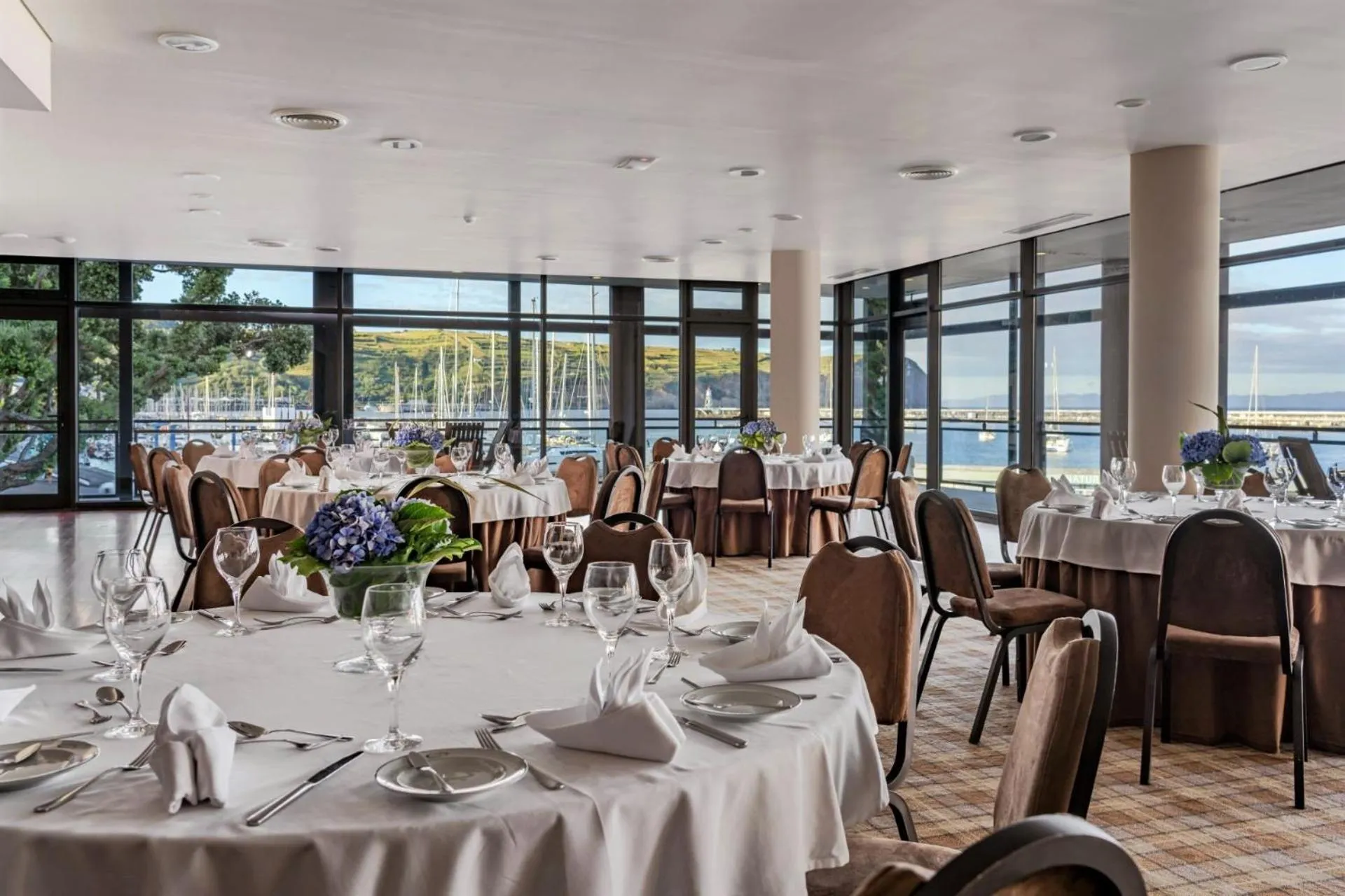 Une salle lumineuse avec des tables dressées pour un repas face à une vue sur la mer et un port de plaisance.