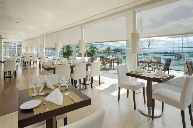 Une salle lumineuse avec des tables dressées pour un repas face à une vue sur la mer et un port de plaisance.