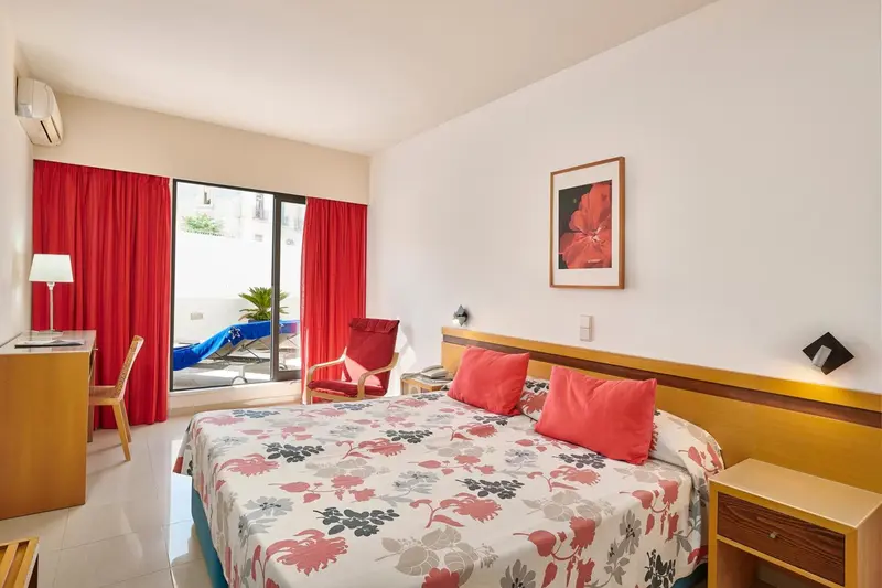 Chambre lumineuse avec un lit recouvert d'une courtepointe fleurie et un fauteuil rouge.