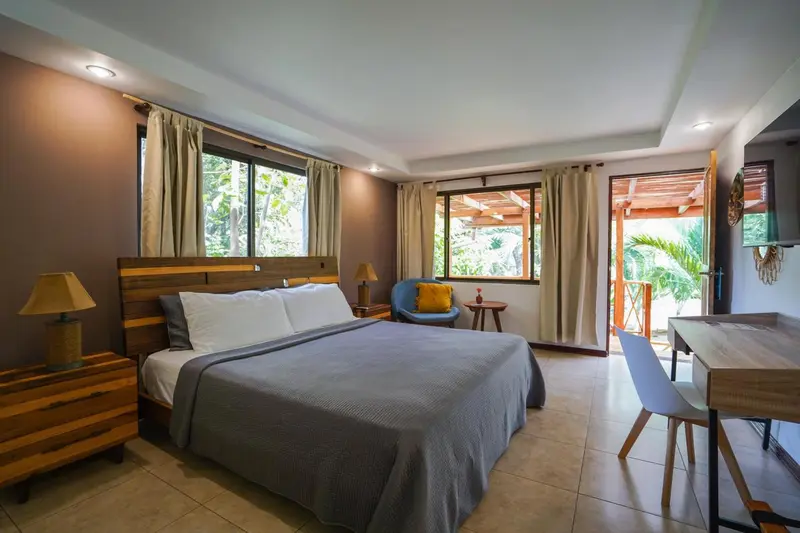 Une chambre lumineuse avec un grand lit et un bureau près d'une porte ouverte sur une terrasse.