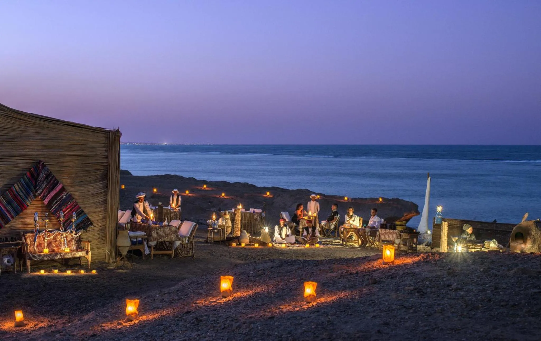 Dîner en plein air sur une plage au coucher du soleil, avec des tables éclairées aux chandelles.