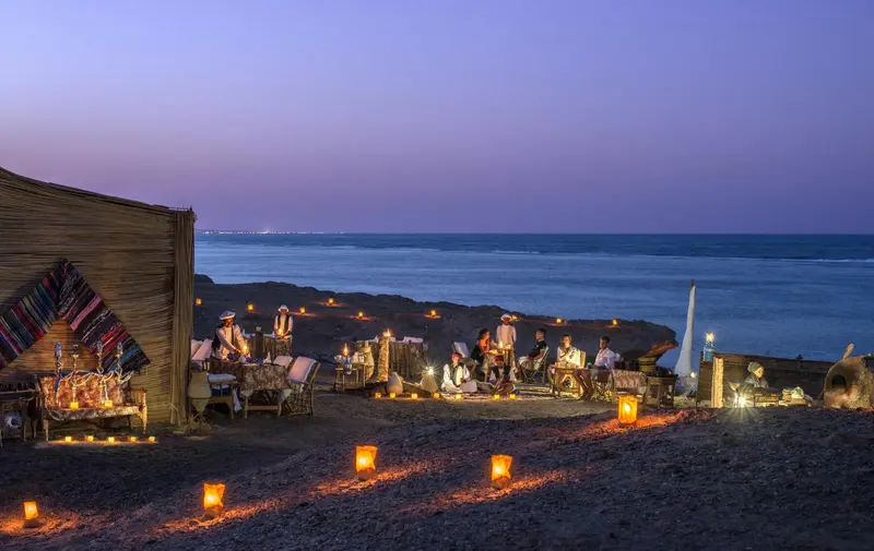 Dîner en plein air sur une plage au coucher du soleil, avec des tables éclairées aux chandelles.