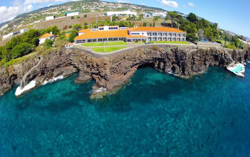 Vue aérienne d'un bâtiment blanc surplombant une côte rocheuse et une mer turquoise.