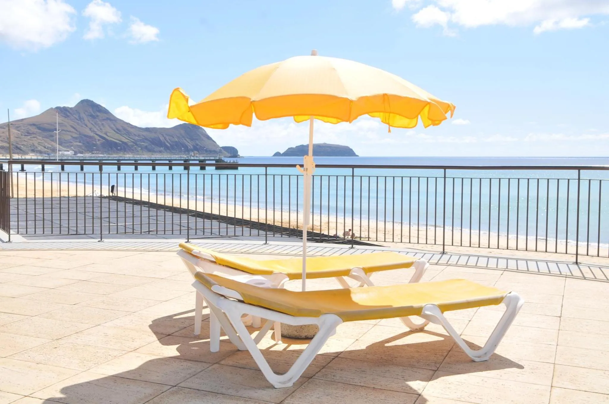 Transats jaunes et parasol sur une terrasse ensoleillée avec vue sur la mer et les montagnes.