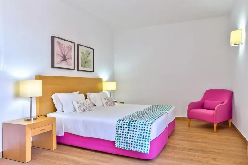 Chambre lumineuse avec un lit double et un fauteuil rose.