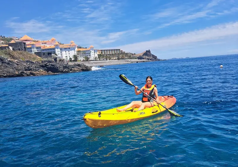 Une personne pratique le kayak en mer face à un littoral rocheux et un bâtiment.