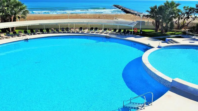 Vue aérienne d'une piscine turquoise entourée de transats et de palmiers.