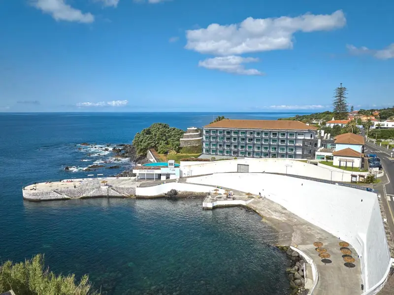 Bâtiment blanc avec toiture brune surplombant une côte rocheuse et une piscine.