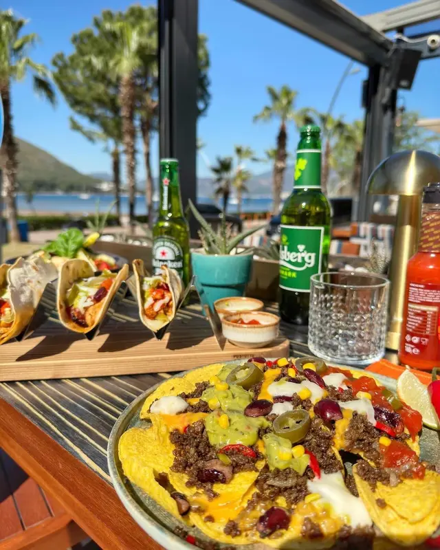 Assiette de tacos et de nachos accompagnée de bières et de sauces sur une table en terrasse.