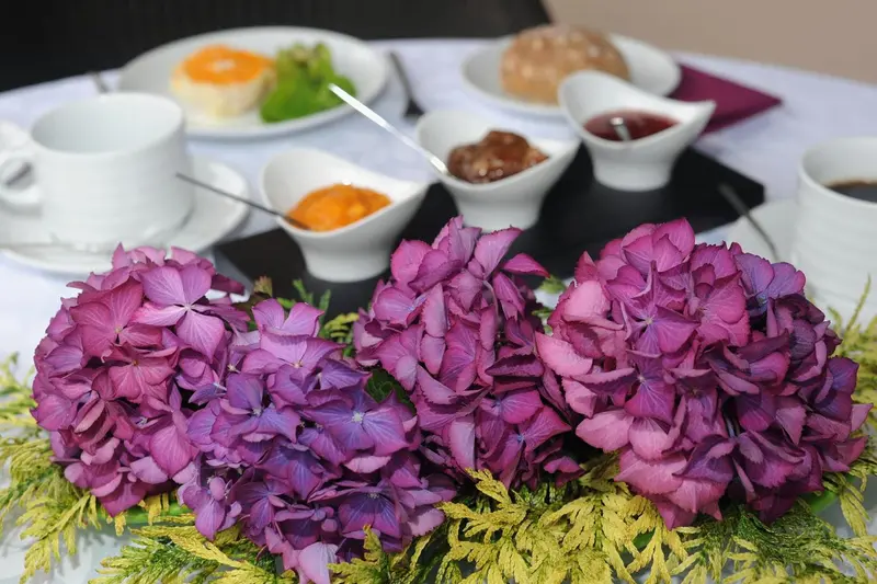 Une table dressée présente un petit-déjeuner avec des viennoiseries, des confitures et une tasse de café, agrémenté d'un bouquet d'hortensias.