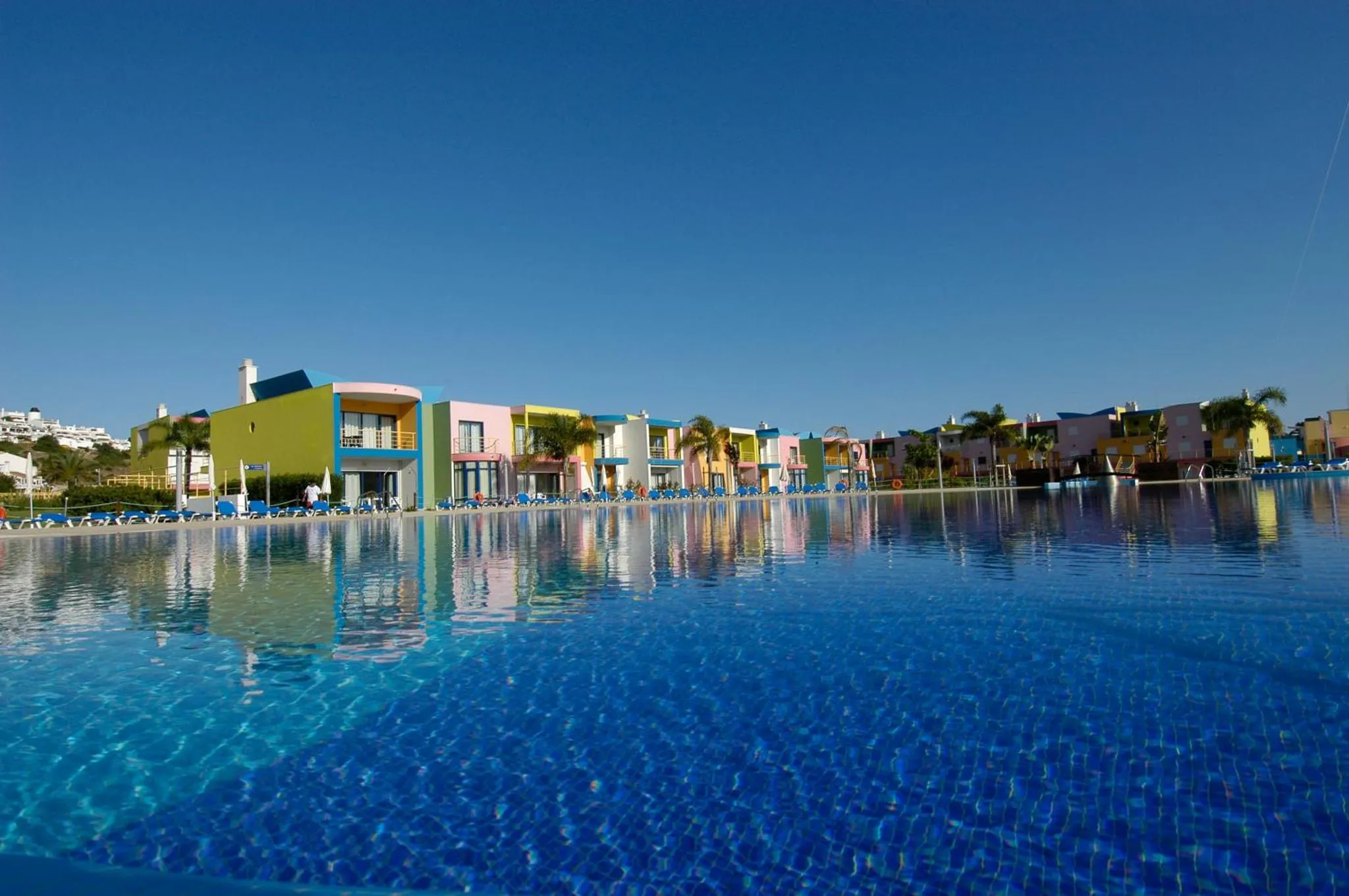 Vue d'une piscine turquoise bordée de transats devant des bâtiments colorés.