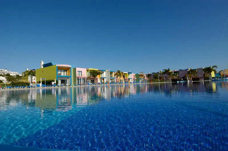 Vue d'une piscine turquoise bordée de transats devant des bâtiments colorés.