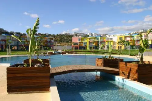 Vue d'une piscine avec un ponton en bois et des bâtiments colorés en arrière-plan.