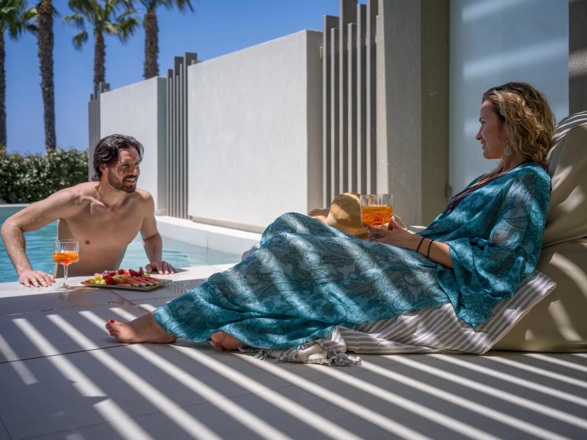 Une femme se détend sur un transat au bord d'une piscine, un homme la rejoint avec des boissons.