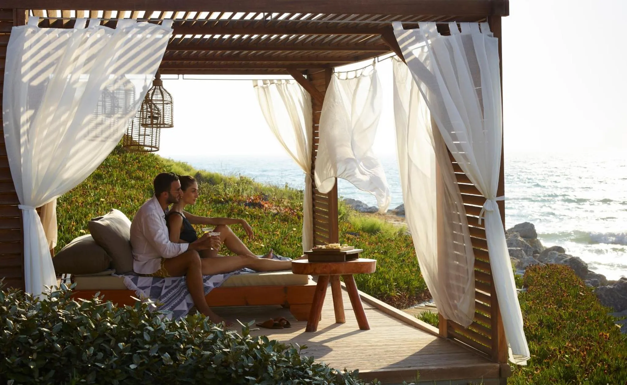 Un couple se détend sur une plateforme en bois ombragée face à la mer.