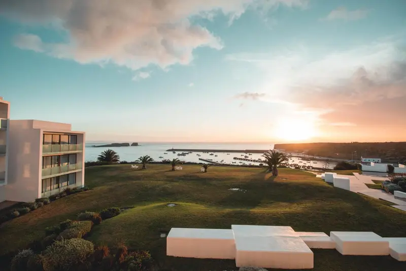 Vue d'un bâtiment blanc avec des balcons donnant sur une pelouse verdoyante et la mer au coucher du soleil.