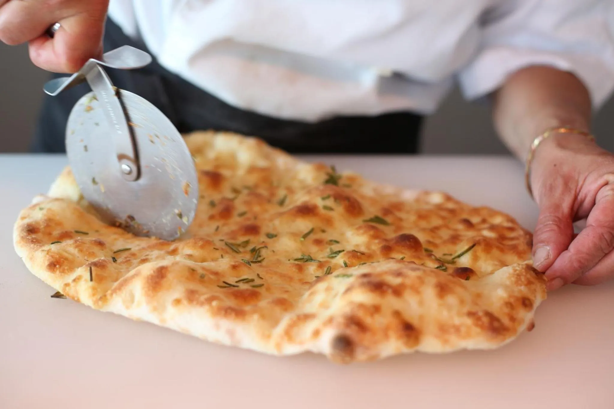 Une personne coupe une grande focaccia au fromage et aux herbes sur une surface blanche.