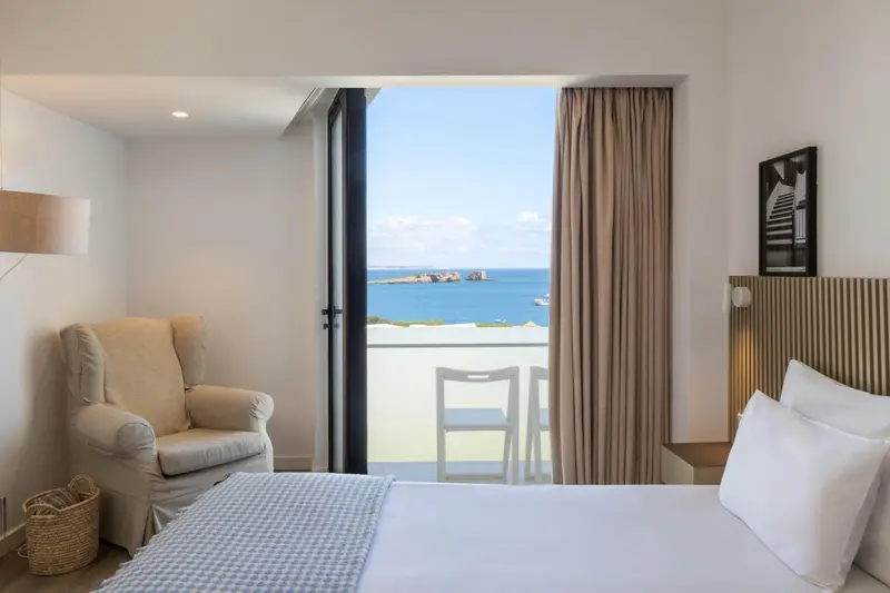 Une chambre lumineuse avec un lit fait et un fauteuil près d'une baie vitrée donnant sur la mer.