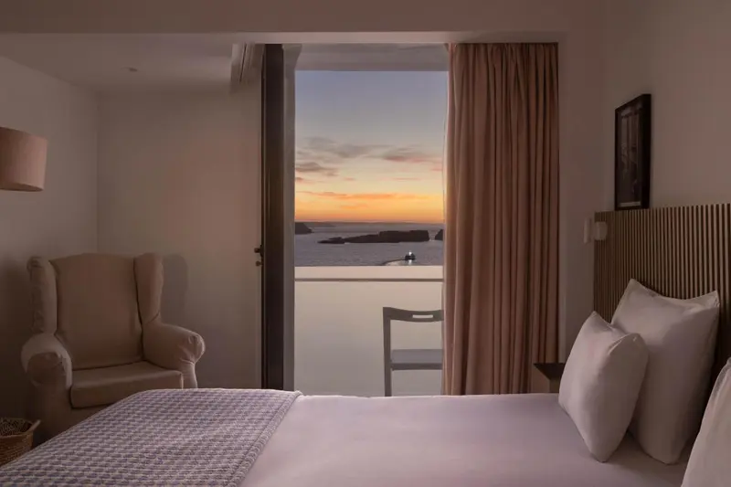 Une chambre avec un lit et un fauteuil donnant sur une vue mer au coucher du soleil.