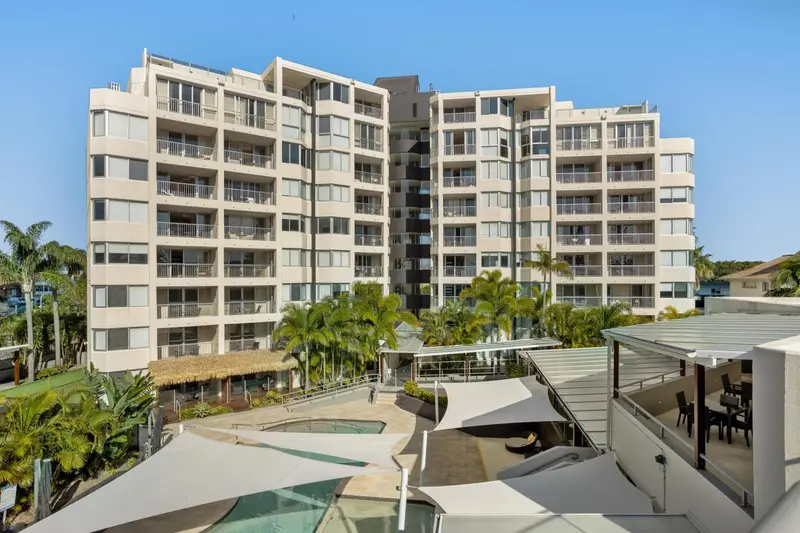 Bâtiment beige à plusieurs étages avec des balcons et une piscine en contrebas.