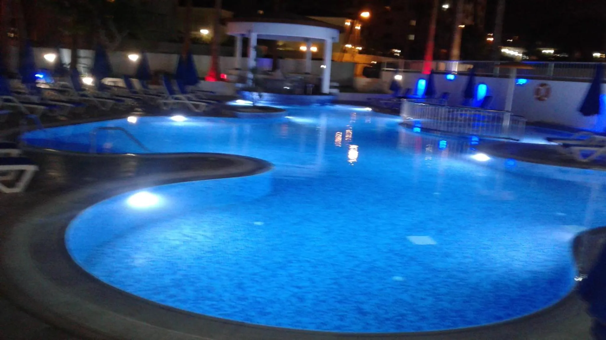 Piscine illuminée avec des transats et des palmiers en arrière-plan.
