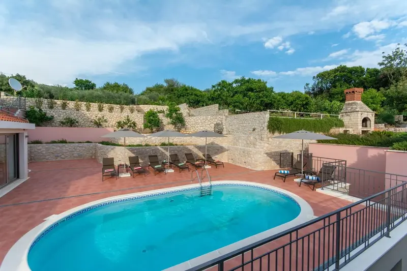 Une piscine rectangulaire avec des transats et des parasols est entourée d'un mur en pierre et de végétation.