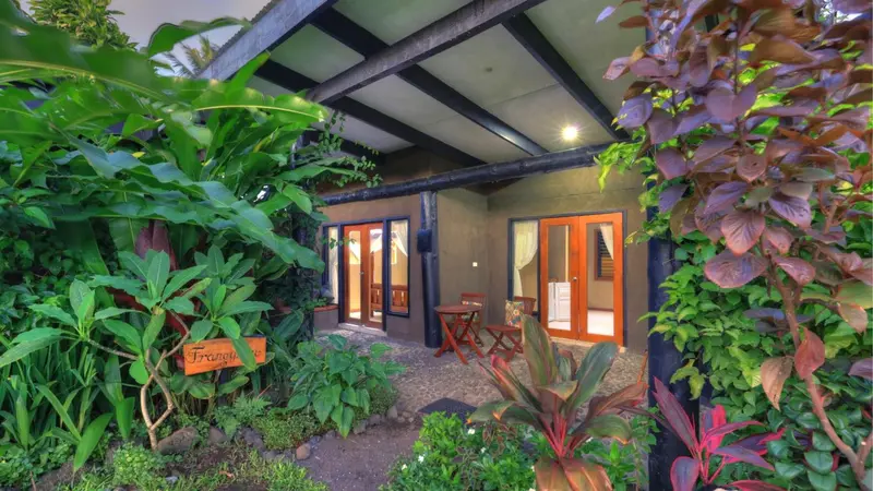 Une terrasse ombragée donne sur une entrée avec une porte orange et des plantes tropicales.