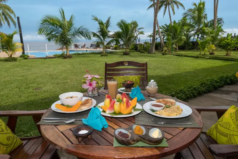 Une table ronde en bois est dressée avec un petit-déjeuner copieux sur une terrasse verdoyante.