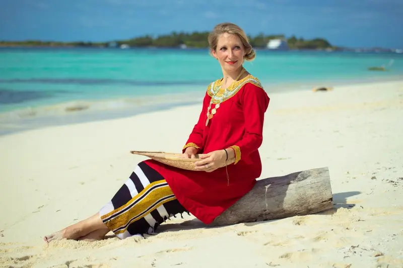 Une femme souriante est assise sur une plage de sable blanc face à une mer turquoise.