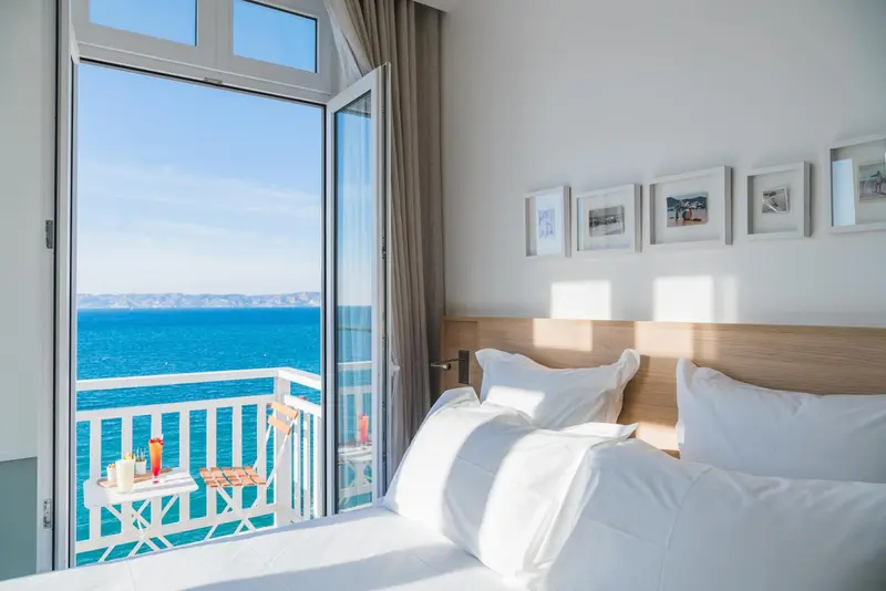 Une chambre lumineuse avec un lit fait et une vue sur la mer depuis un balcon.