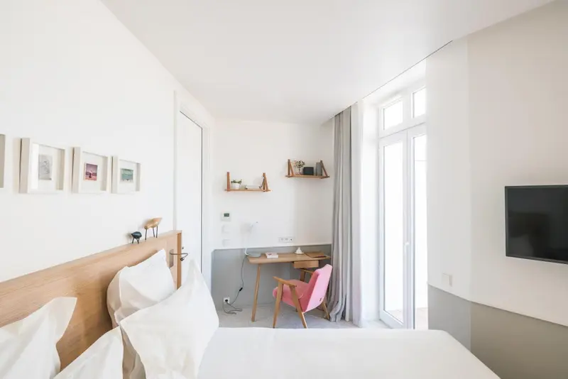 Chambre lumineuse avec un lit blanc et un bureau rose près d'une fenêtre.
