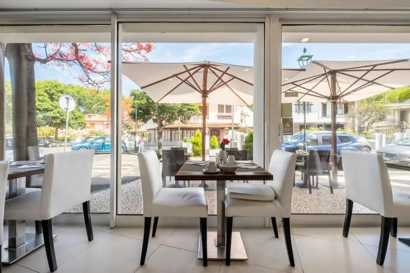 Tables et chaises blanches disposées devant de grandes baies vitrées donnant sur une terrasse ombragée.