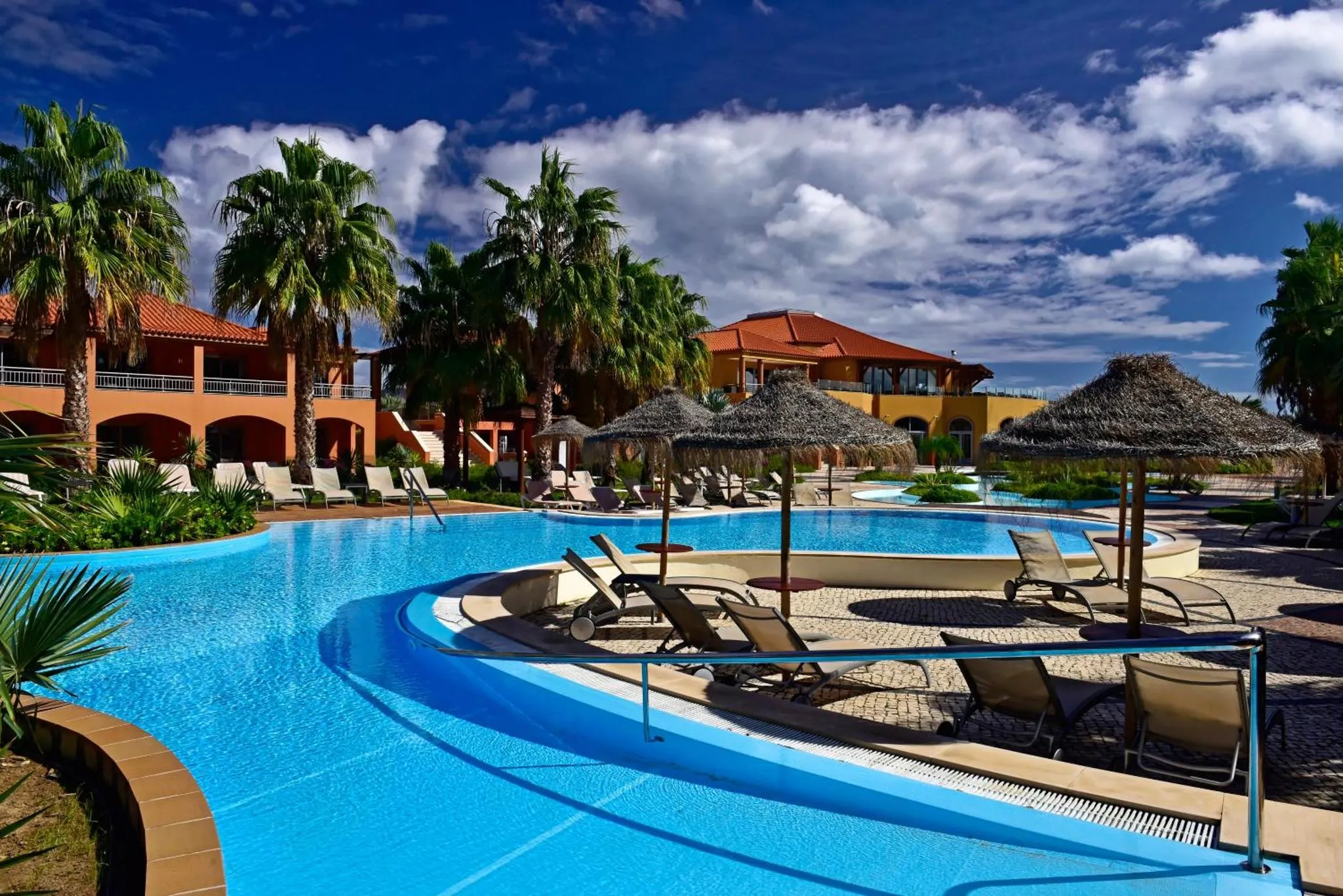 Une piscine turquoise bordée de transats et de palmiers sous un ciel bleu.