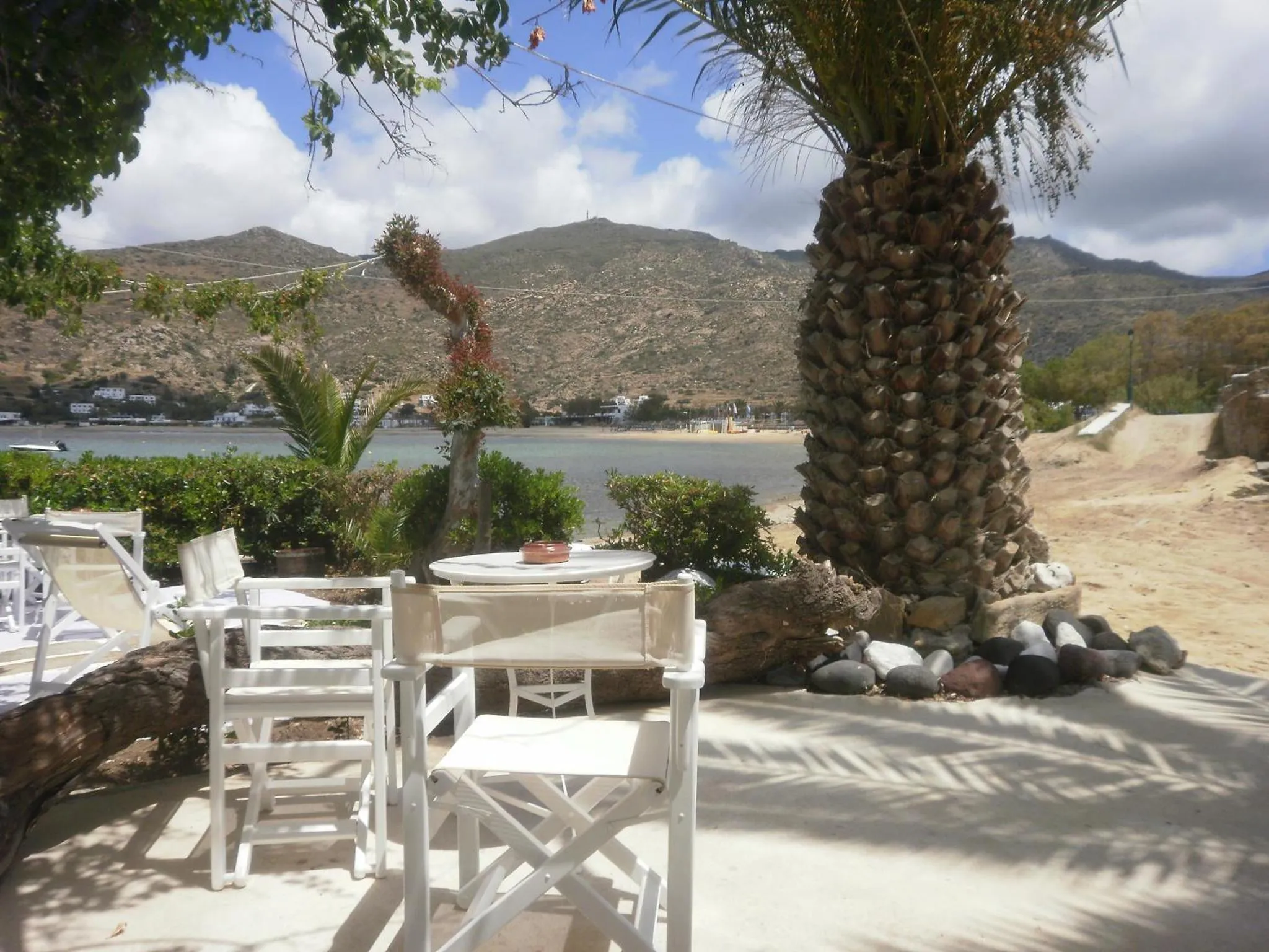 Tables et chaises blanches disposées sur une terrasse ensoleillée avec vue sur la mer et les montagnes.