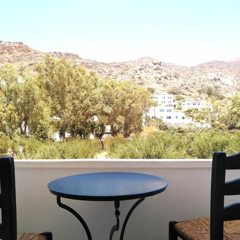 Une table ronde et deux chaises en osier se trouvent sur une terrasse ensoleillée avec vue sur un paysage montagneux.