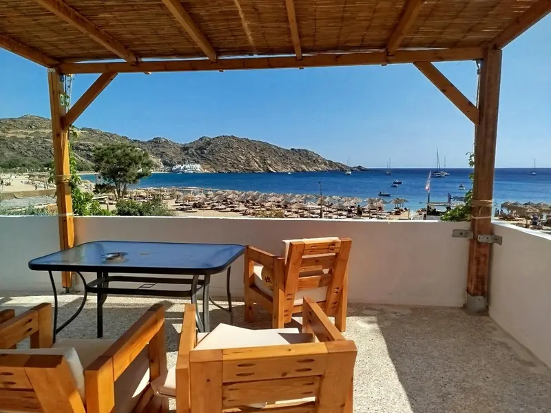 Une table et des chaises en bois sont disposées sur une terrasse ombragée avec vue sur une plage.