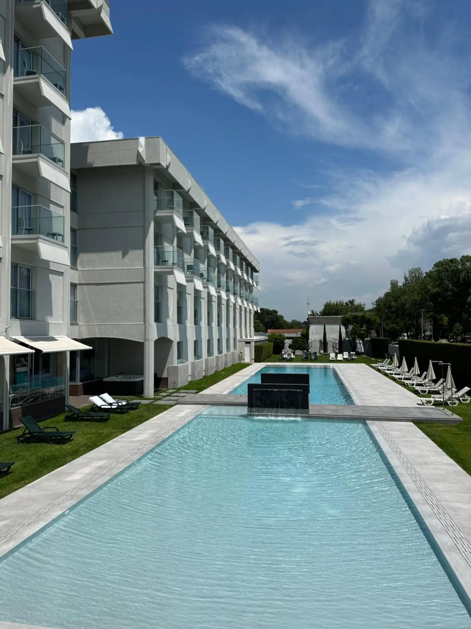 Une longue piscine rectangulaire bordée de transats et de végétation luxuriante devant un bâtiment moderne.